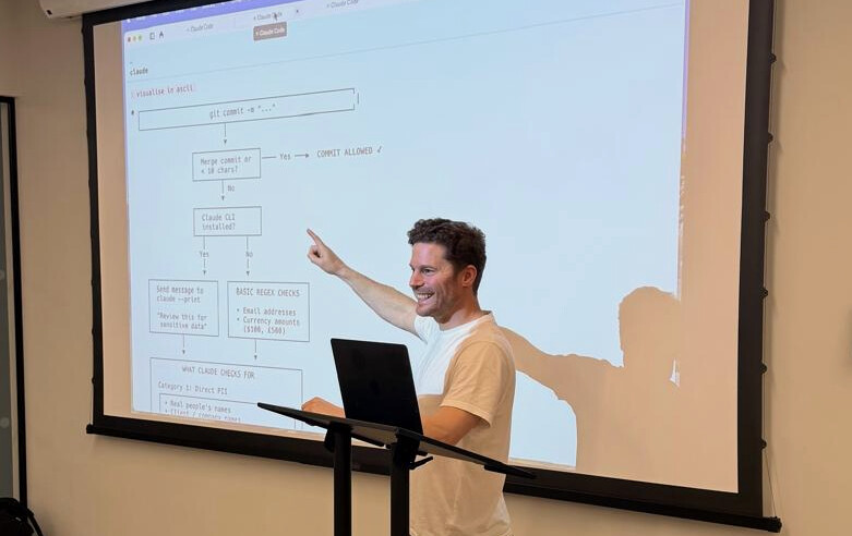 Alex Appelbe presenting at an event for AI leaders in London, standing at a podium and pointing at a large projected screen. The screen shows a flowchart of a commit-time check that scans for sensitive data – names, email addresses, currency amounts – before anything is saved.